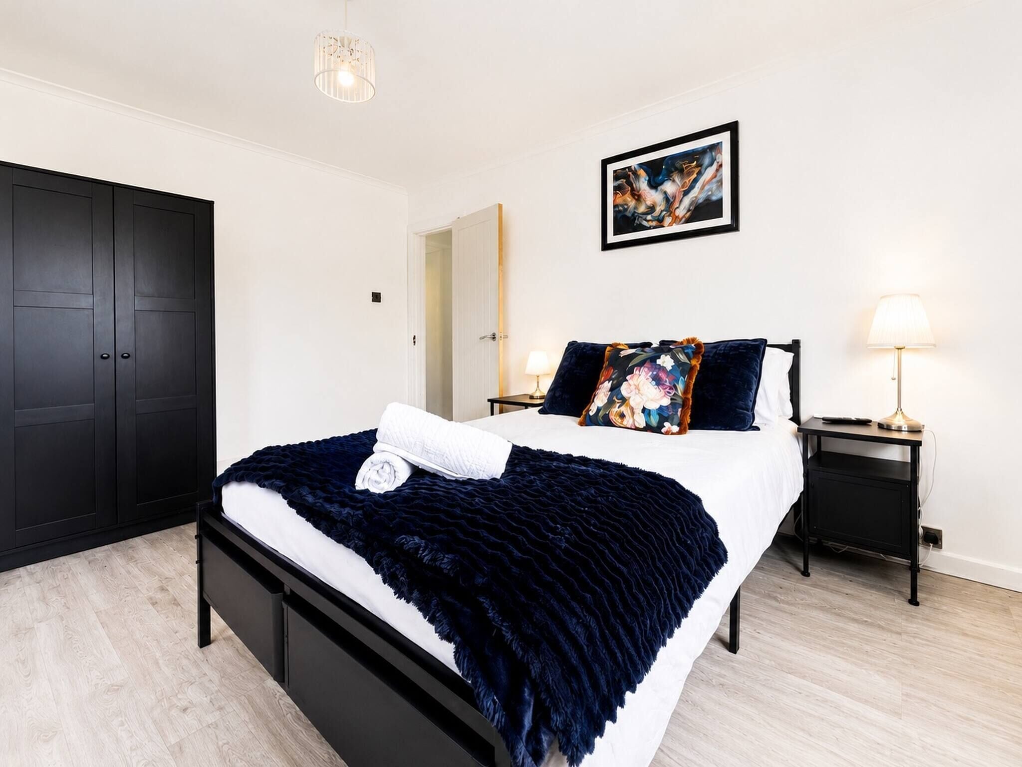 A modern bedroom with a large bed, a wardrobe, a bedside table with a lamp, and a painting on the wall.