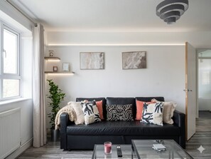 A modern living room with a black sofa, a glass coffee table, and a potted plant. - Ultra-spacious 2 BR In Central Wimbledon! (London)