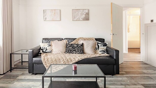 En moderne stue med en sort lædersofa, to indrammede abstrakte malerier, et sofabord i glas og en dør, der fører til et andet rum.