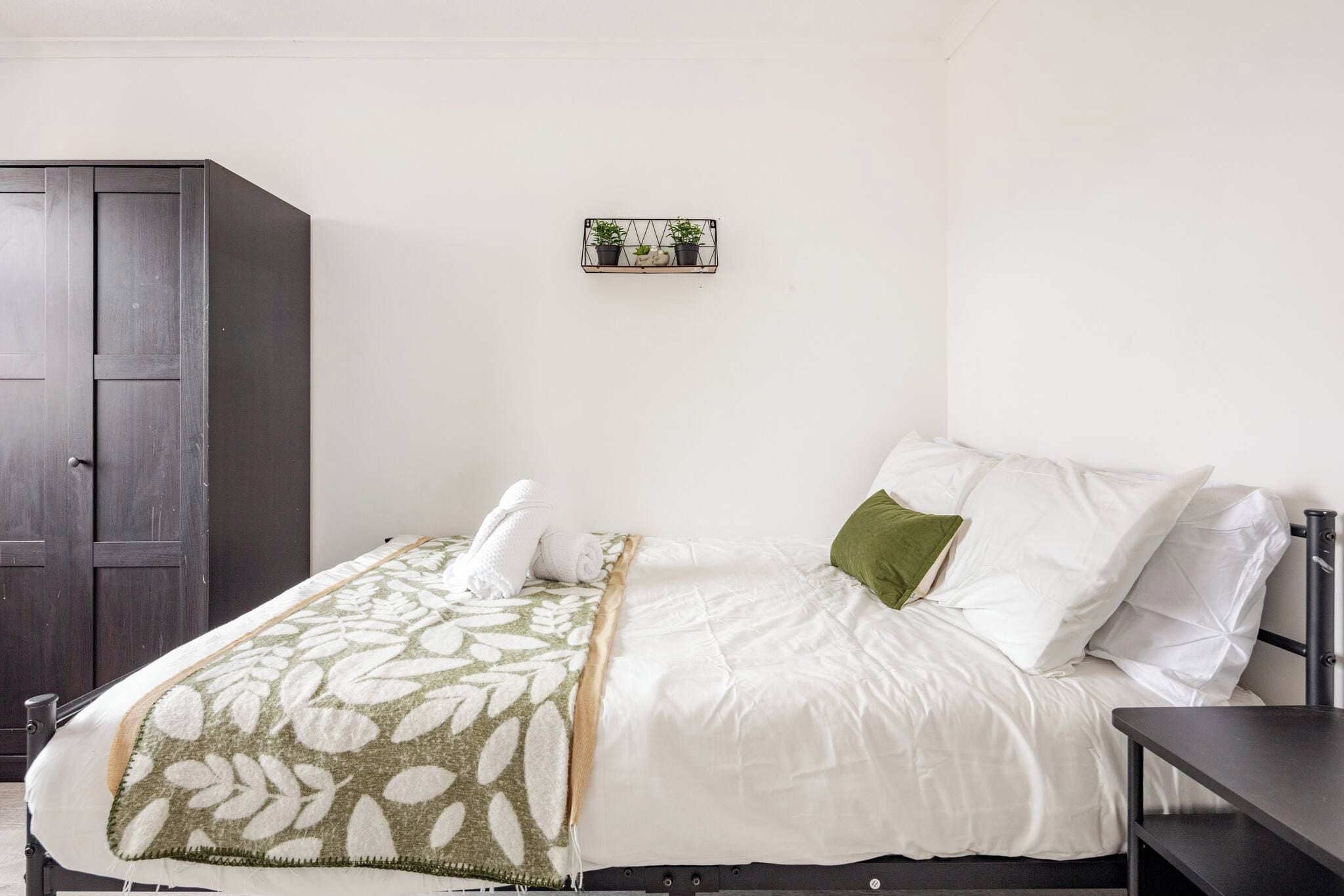 Un lit bien fait, recouvert d’une couverture à motifs blancs et verts, avec un oreiller vert et une petite étagère ornée de plantes accrochée au mur.