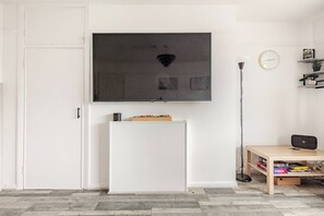Ein Zimmer mit einem an der Wand befestigten Flachbildfernseher, einem weißen Schrank darunter, einem Holztisch mit einem Laptop und einer Stehlampe.