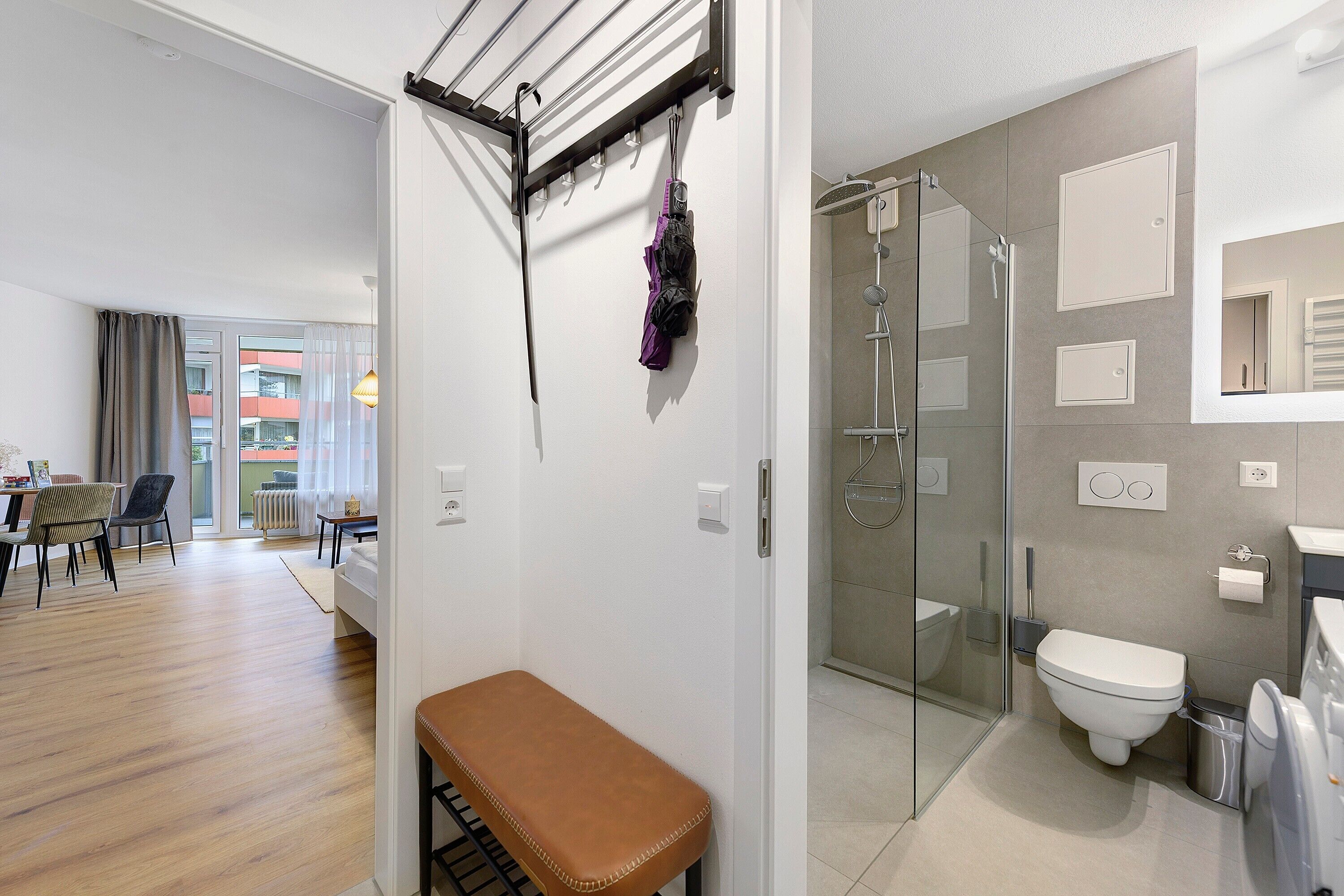 Ein modernes Badezimmer mit Duschkabine aus Glas, Toilette und Waschbecken. Im angrenzenden Wohnbereich befindet sich eine Sitzbank.