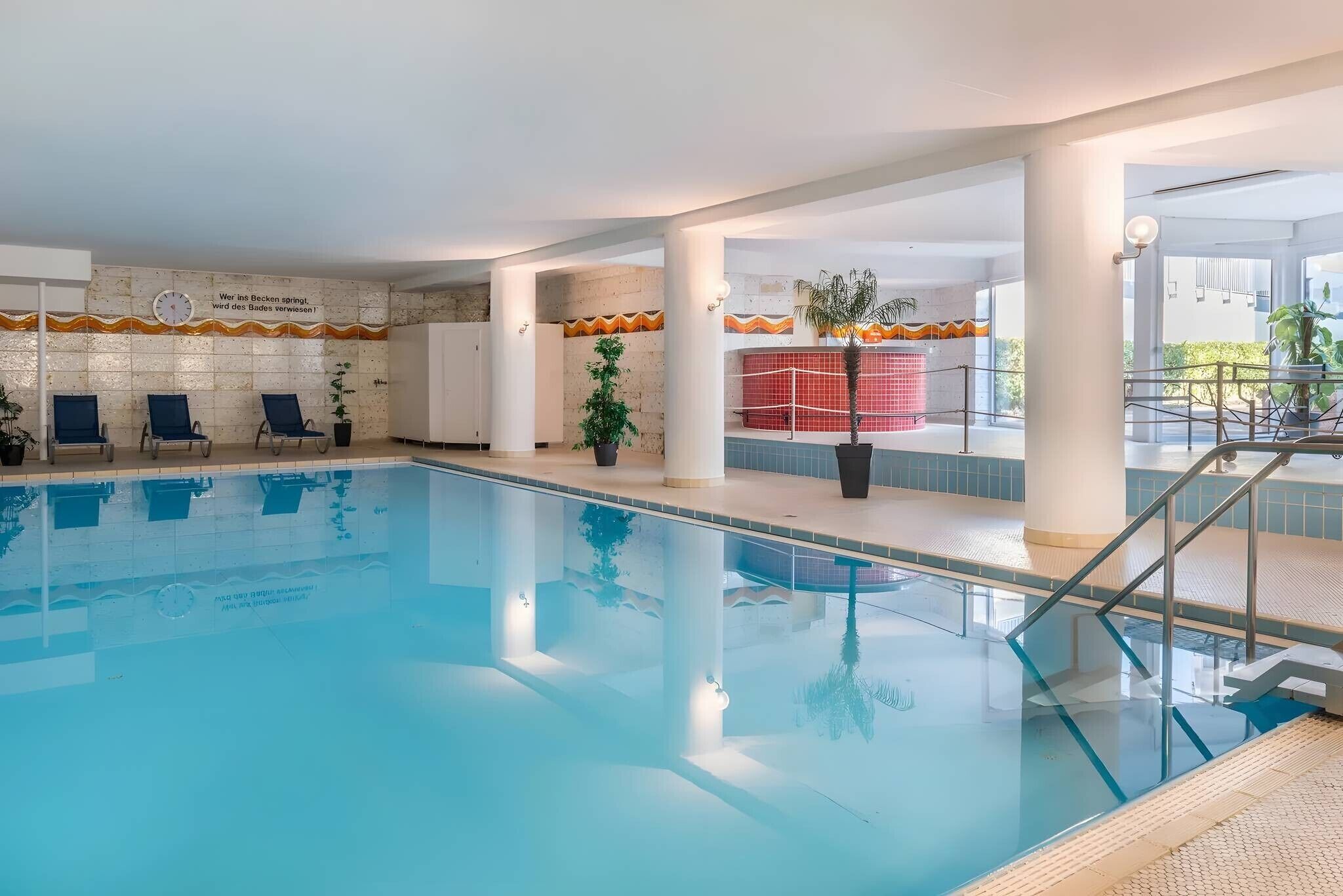 An indoor swimming pool with clear water, surrounded by lounge chairs and potted plants.