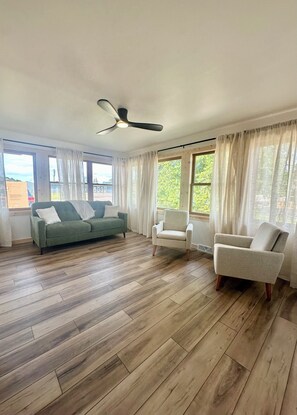 Un amplio salón con suelo de madera, un ventilador de techo y ventanas grandes con cortinas.