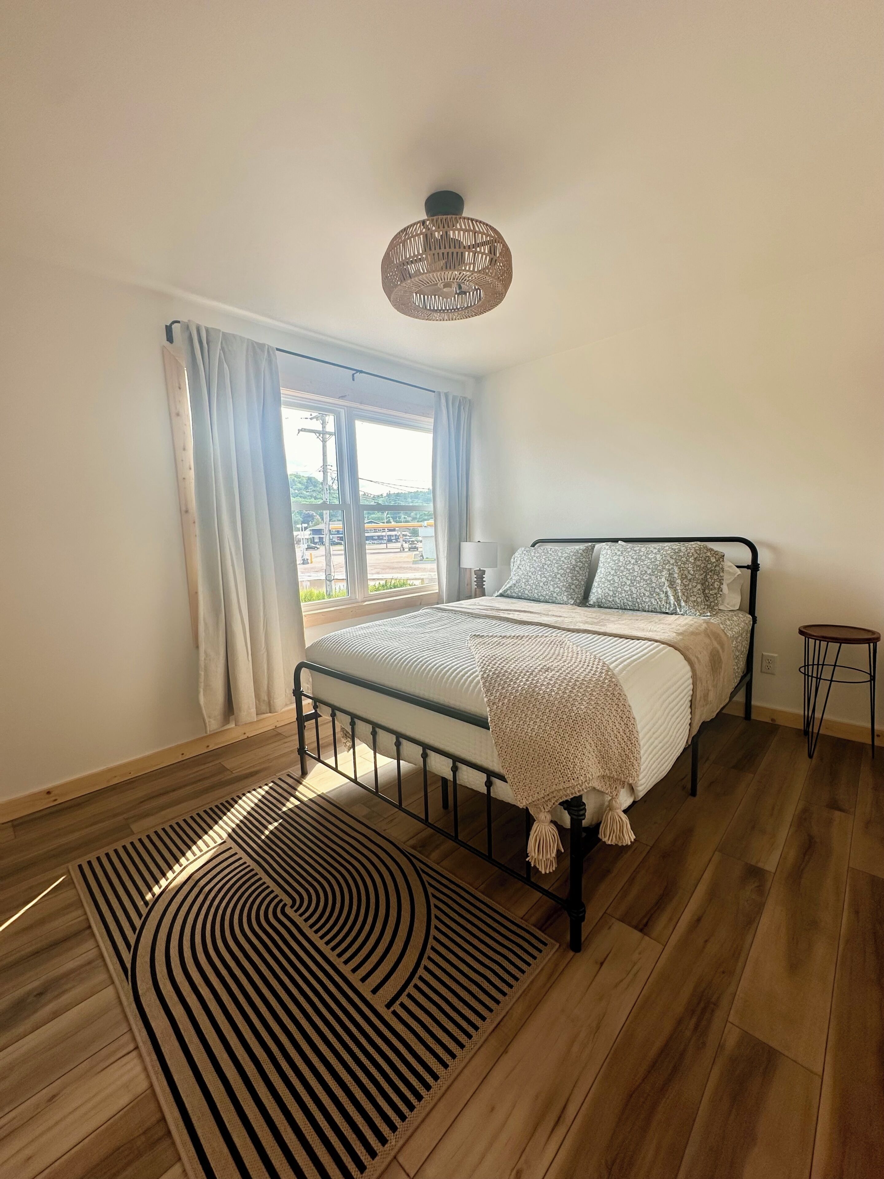 A bedroom with a bed, a bedside table, a lamp, a hanging light fixture, a window with curtains, and a patterned rug.