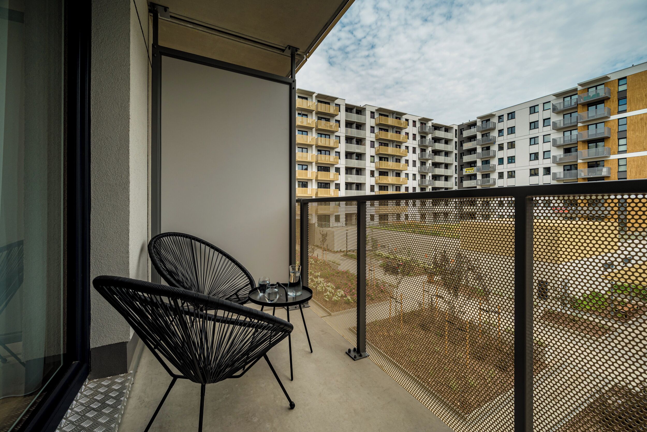 Un balcon avec une chaise noire et une petite table, donnant sur un immeuble d’habitation doté de balcons.