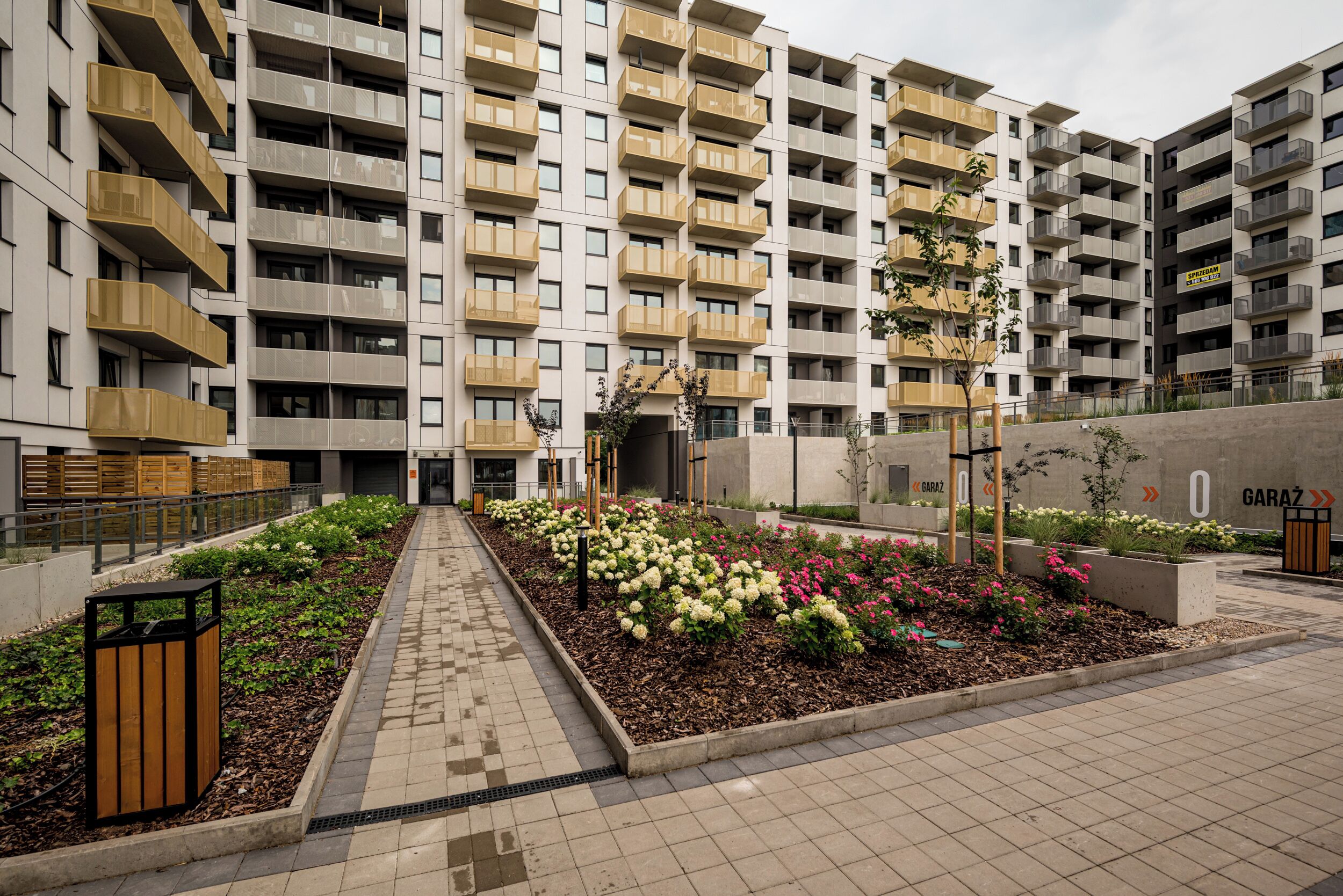 A modern apartment building with multiple balconies, a central garden area with flowers, and a paved walkway.