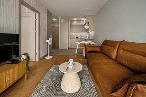 A modern living room with a brown sofa, a TV on a wooden cabinet, a small table with a white fan, and a kitchen in the background. - RentPlanet - Apartament Braterska (Wrocław)