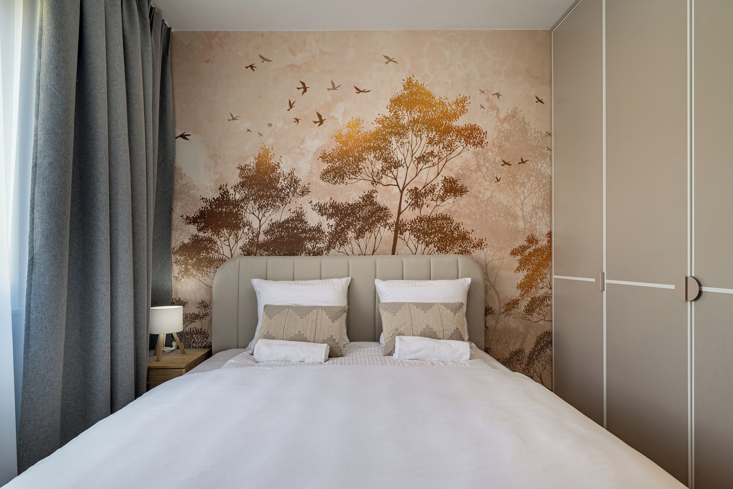 A bedroom with a bed, pillows, a nightstand, a lamp, and a wall with a nature-themed mural.
