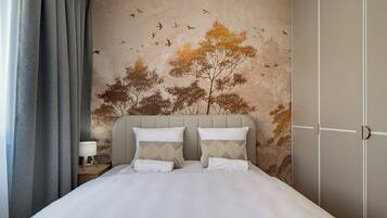 A bedroom with a bed, pillows, a nightstand, a lamp, and a wall with a nature-themed mural.