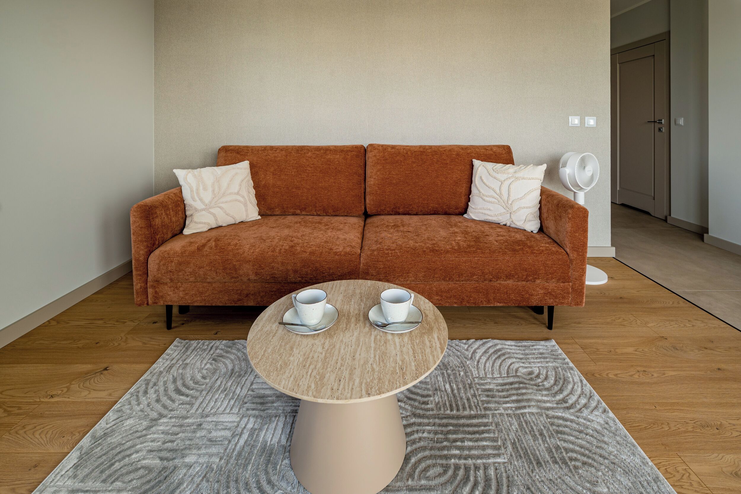 Ein modernes Wohnzimmer mit einem braunen Sofa, zwei weißen Kissen, einem runden Couchtisch mit zwei weißen Tassen und Untertellern sowie einem weißen Ventilator.