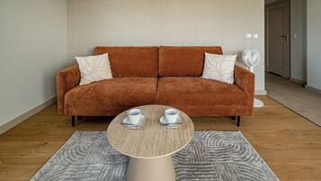 Ein modernes Wohnzimmer mit einem braunen Sofa, zwei weißen Kissen, einem runden Couchtisch mit zwei weißen Tassen und Untertellern sowie einem weißen Ventilator.