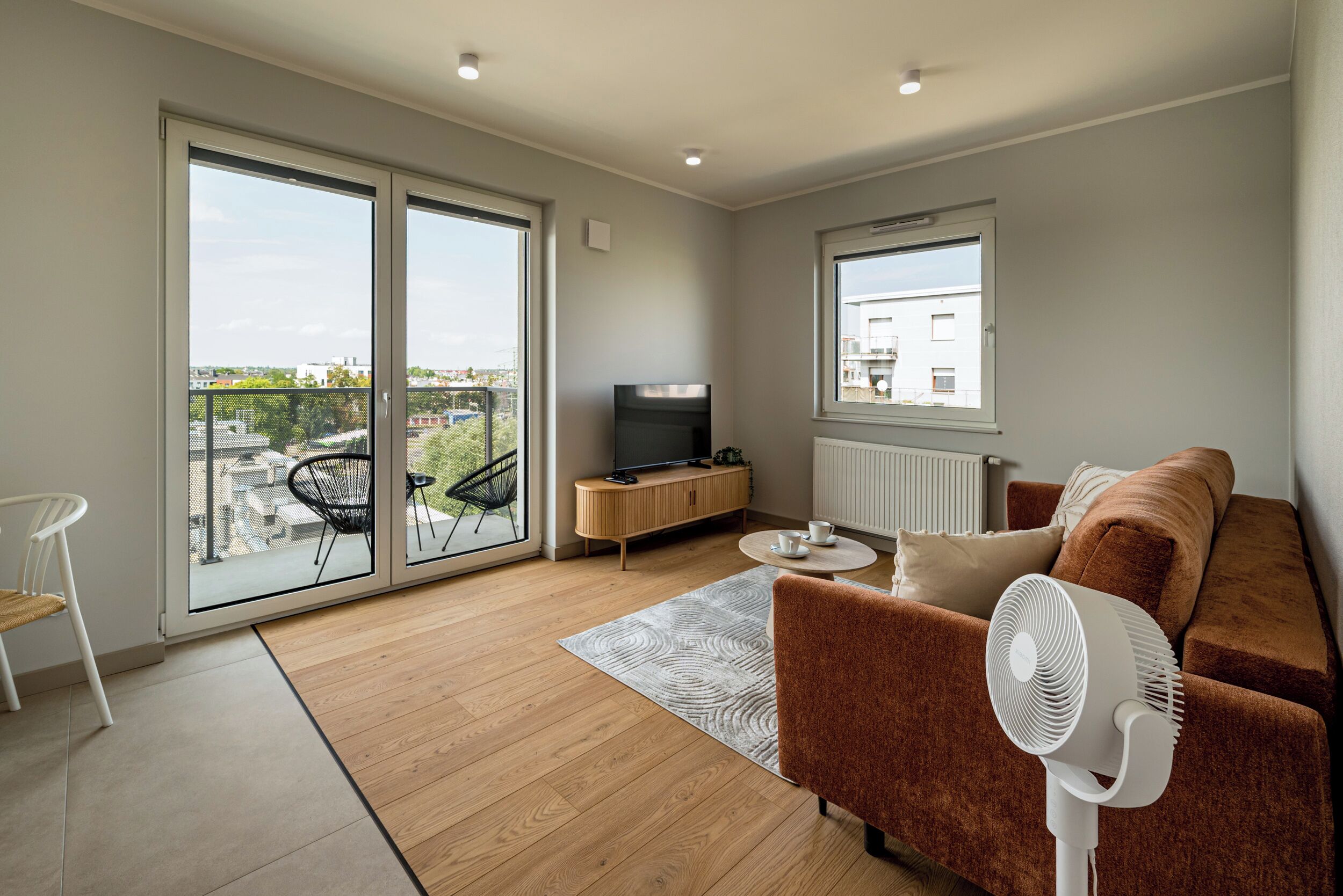 A modern living room with a sofa, a TV, a fan, and a view of the city through large windows.