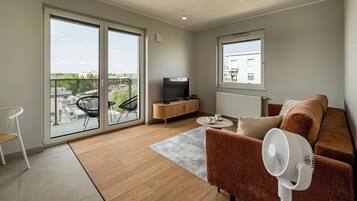 A modern living room with a sofa, a TV, a fan, and a view of the city through large windows.