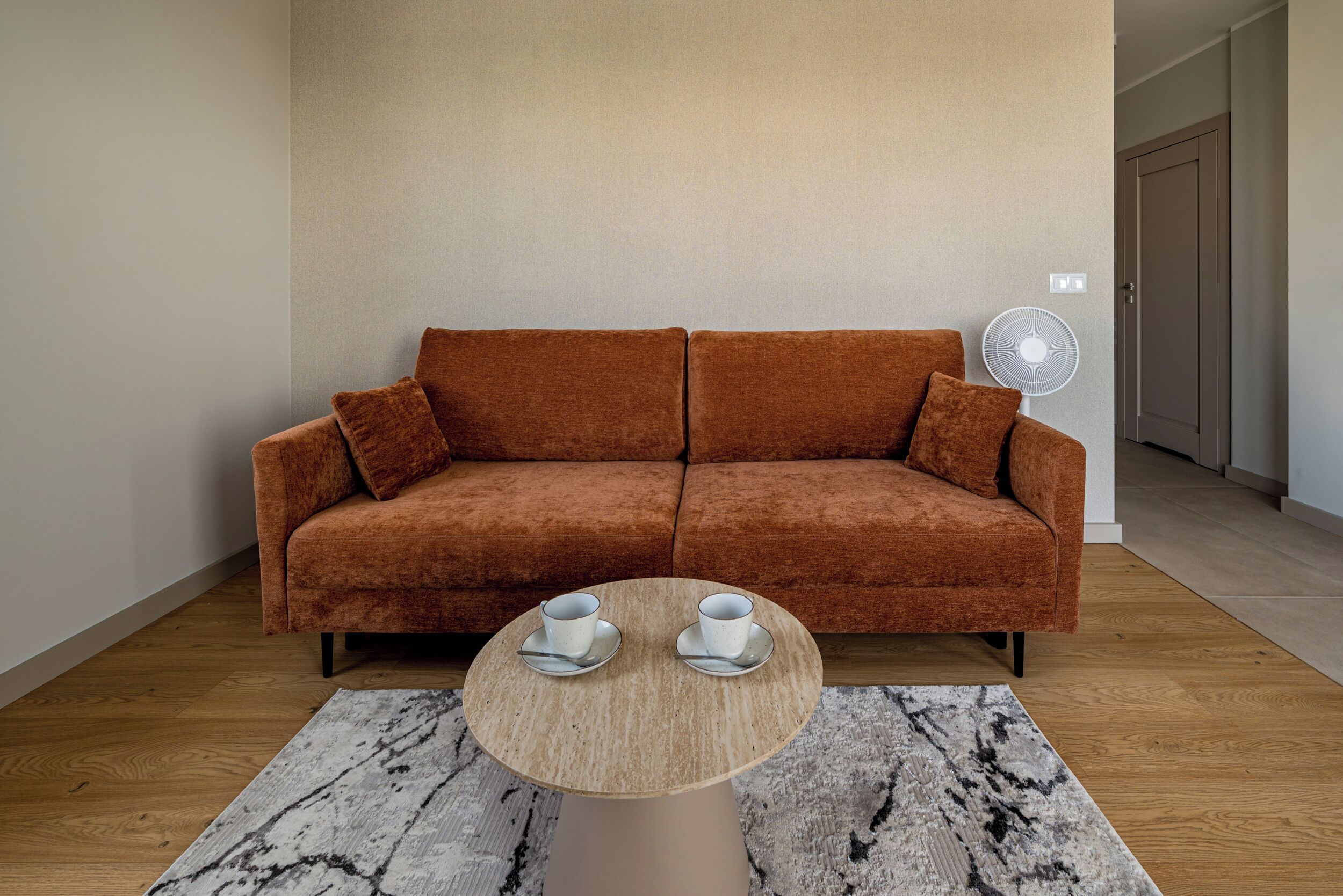 Una habitación con un sofá marrón, una mesa redonda con dos tazas y un ventilador en la pared.