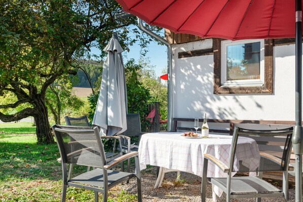 An outdoor seating area with a table and chairs, a red umbrella, and a view of a tree and building in the background. - Vacation home 'am quittenbaum', family vacation on Lake Constance (Überlingen)