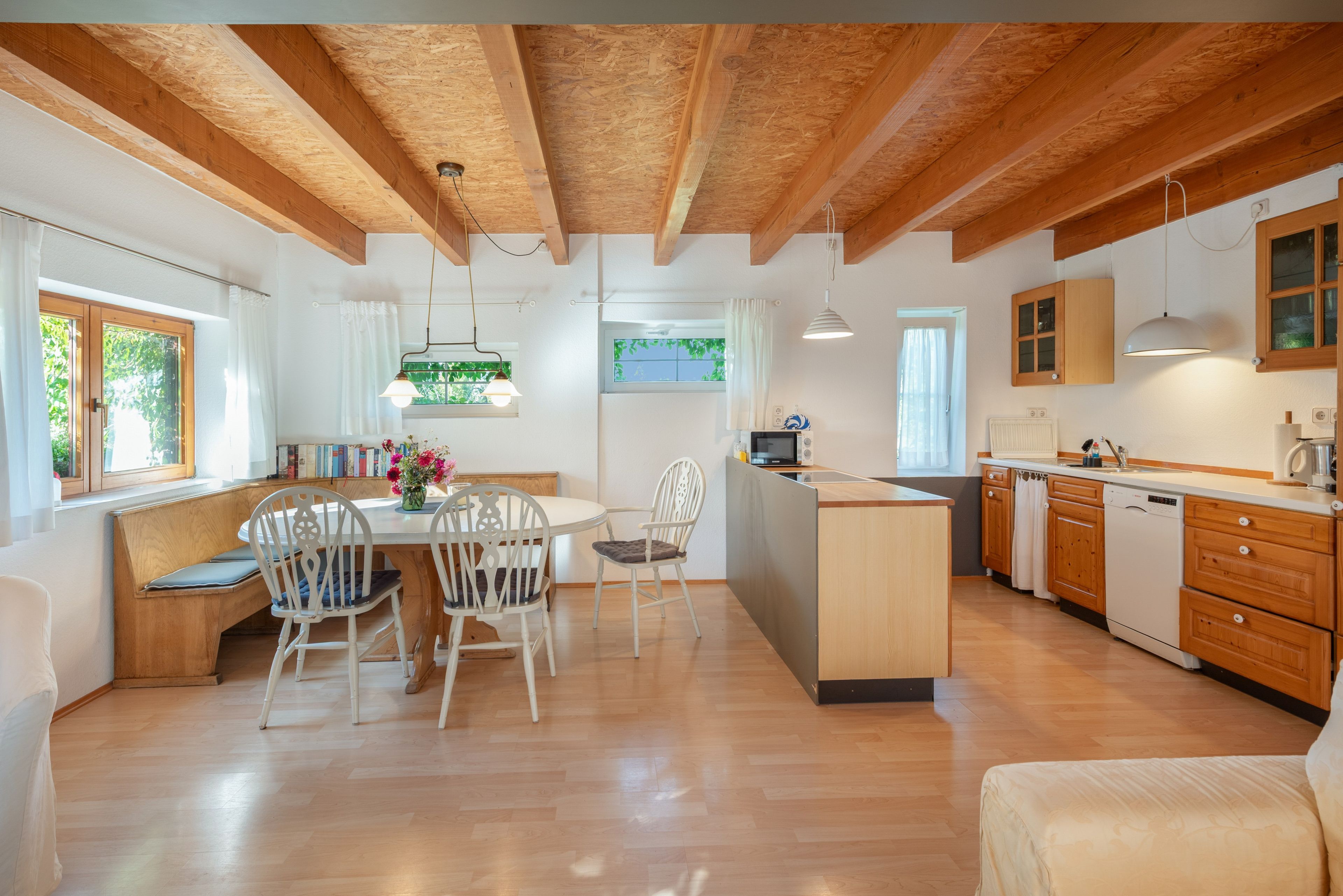 A modern kitchen with wooden beams, a dining area with chairs, and a view of greenery outside.