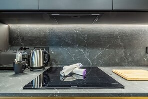 Eine Küche mit einer schwarzen Induktionskochplatte, einem Wasserkocher, einem Toaster und einem Schneidebrett auf einer Marmorarbeitsplatte.