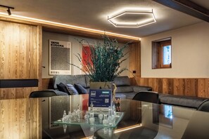Ein modernes Interieur mit einem Glastisch, einem Schachspiel, Wandpaneelen aus Holz und einer geometrischen Leuchte.