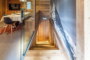 Un interior moderno con una escalera de madera que baja a un nivel inferior, una zona de comedor con mesa y sillas, y un cuadro grande en la pared.