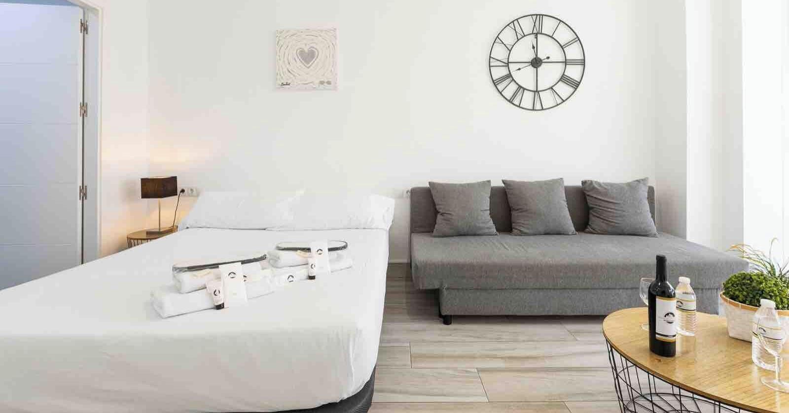 Habitación de hotel con una cama, un sofá, un reloj de pared, una mesita con una botella y vasos, y una obra de arte en la pared.
