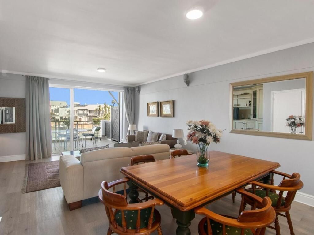 A spacious living area with a wooden dining table, green upholstered chairs, a sofa, and a large window offering a view of the outside.