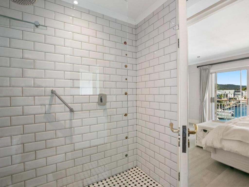 Habitación de hotel con ducha de azulejos, cama con ropa blanca y vista a un puerto deportivo a través de un ventanal grande.
