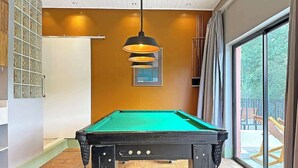 Una mesa de billar en una habitación con un ventanal grande, suelo de madera y una lámpara colgante.