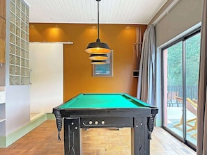 A pool table in a room with a large window, wooden flooring, and a hanging pendant light.
