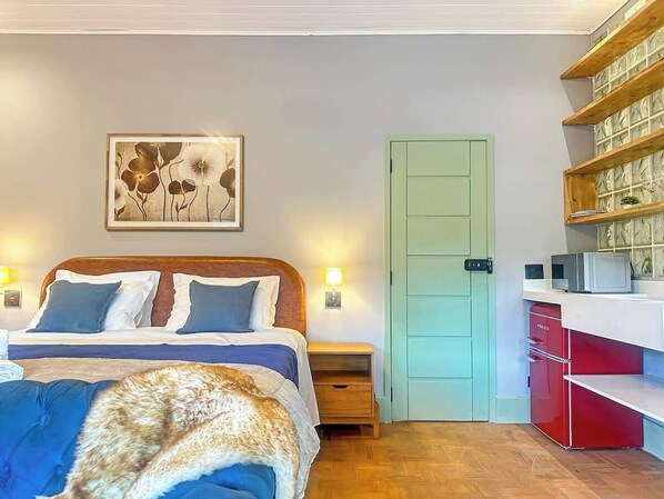 A bedroom with a bed, bedside tables, a door, and a kitchenette area.