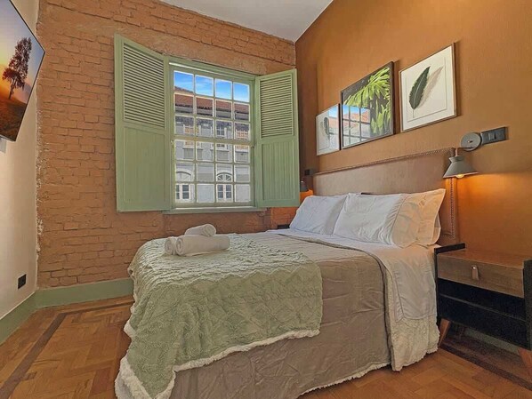 A bedroom with a bed, a nightstand, a lamp, and a window with shutters.