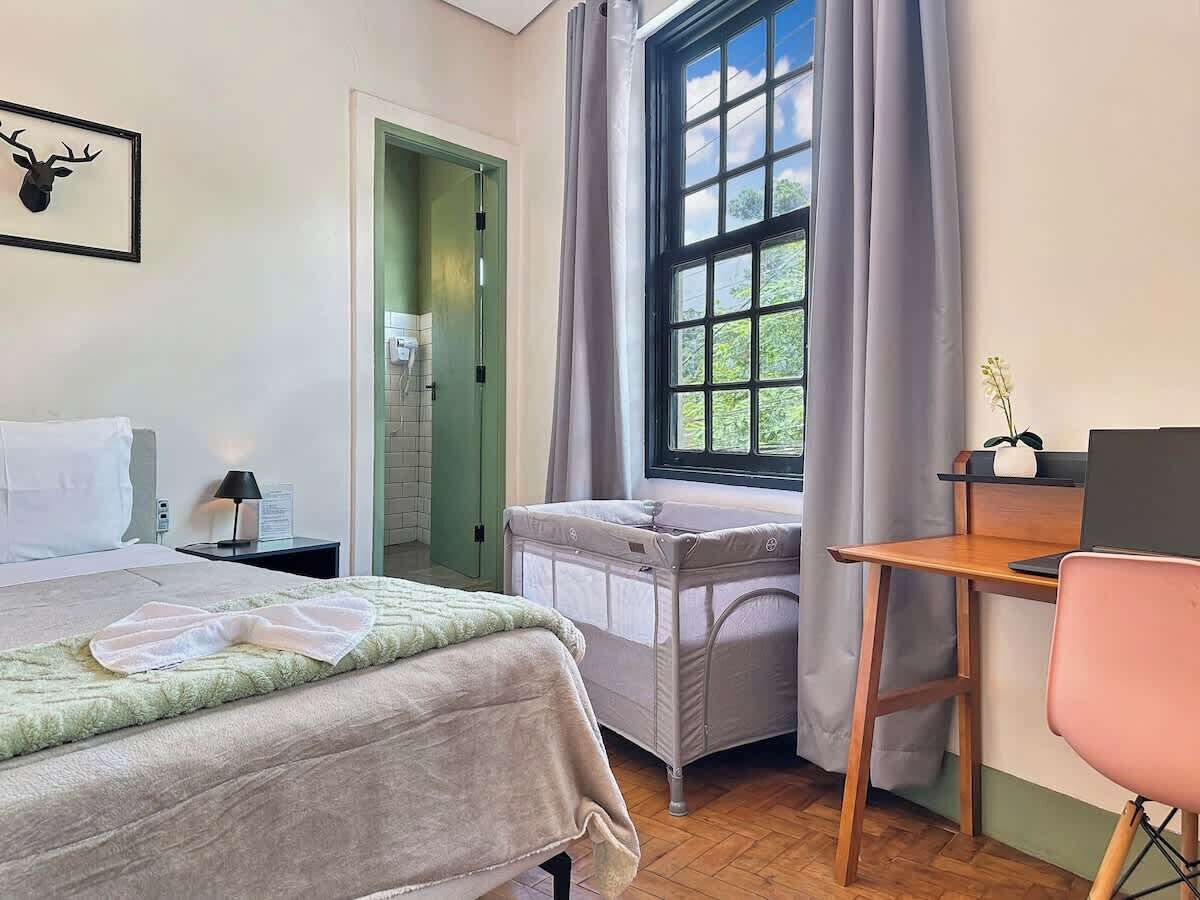 A bedroom with a bed, a desk, a chair, a window with curtains, and a door leading to a bathroom.