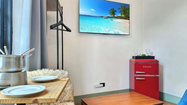 A room with a wooden table, a red Philco filing cabinet, a blue chair, a fluffy beige sofa, and a wall-mounted television displaying a beach scene. - Villa_Aberdeen _01 · Villa Aberdeen | Wi-Fi | Central | Garagem, Qua... (Campos do Jordão)