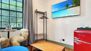 A room with a bed, a small wooden table, a hanging shelf, a red appliance, and a window with a view of a beach. - Villa_Aberdeen _01 · Villa Aberdeen | Wi-Fi | Central | Garagem, Qua... (Campos do Jordão)