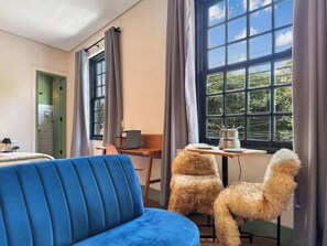 Um quarto com um sofá azul, uma mesa pequena com um laptop, duas poltronas fofas e uma janela com cortinas.