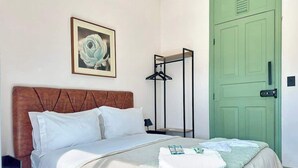 Un lit bien fait, avec une couette blanche et des oreillers, une œuvre d’art encadrée au mur, une porte verte et un porte-manteau noir.