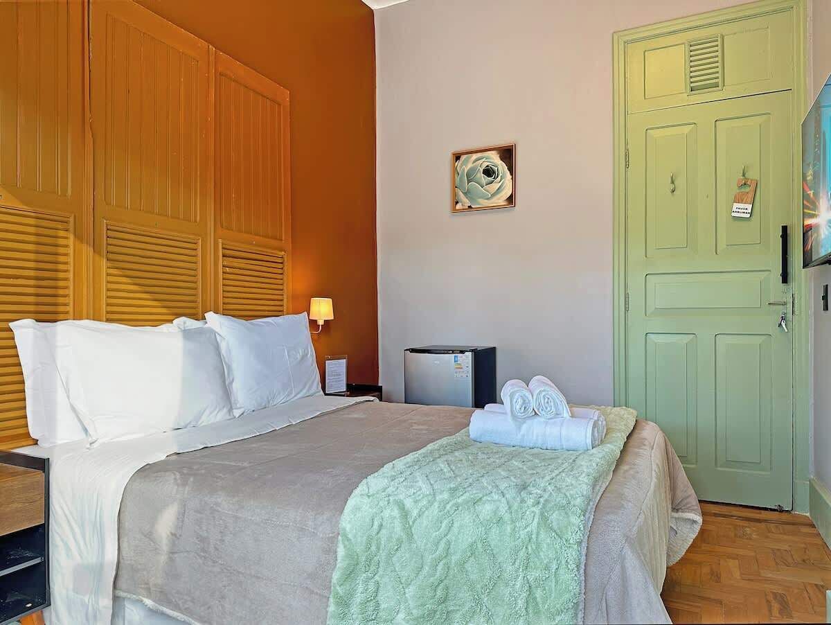 A hotel room with a bed, a nightstand, a lamp, a refrigerator, a green door, and a wall clock.