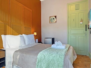 Una habitación de hotel con cama, mesita de noche, lámpara, refrigerador, una puerta verde y un reloj de pared.