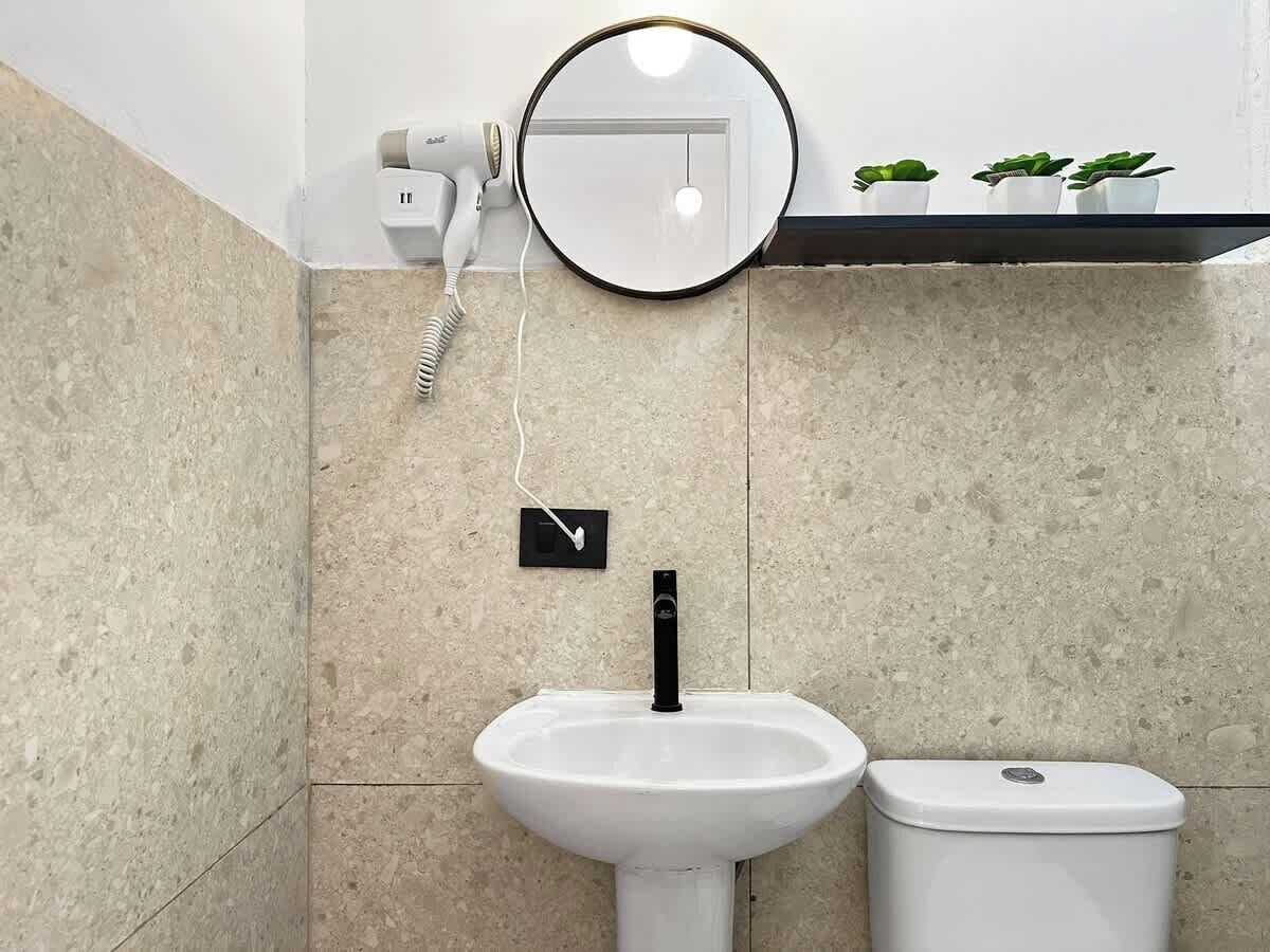 A bathroom with a white pedestal sink, a black faucet, a white toilet, a hairdryer, and a mirror.