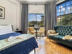 Quarto de hotel com cama, sofá, uma pequena mesa com cadeiras e uma janela grande que oferece vista para o exterior.