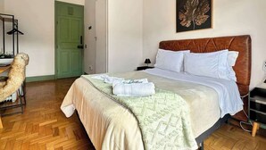 Quarto de hotel com uma cama, uma porta verde, um cabideiro e uma folha emoldurada.