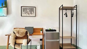 Quarto compacto com uma escrivaninha de madeira, uma cadeira com manta de pele sintética, um refrigerador e uma estante de metal preta.