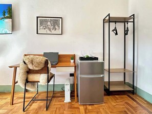Una stanza compatta con una scrivania in legno, una sedia con una coperta in pelliccia, un frigorifero e una scaffalatura in metallo nero.