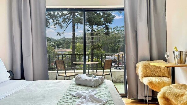 Quarto de hotel com uma cama, uma pequena mesa com uma garrafa e copos, uma poltrona macia e uma varanda com vista para árvores e edifícios.
