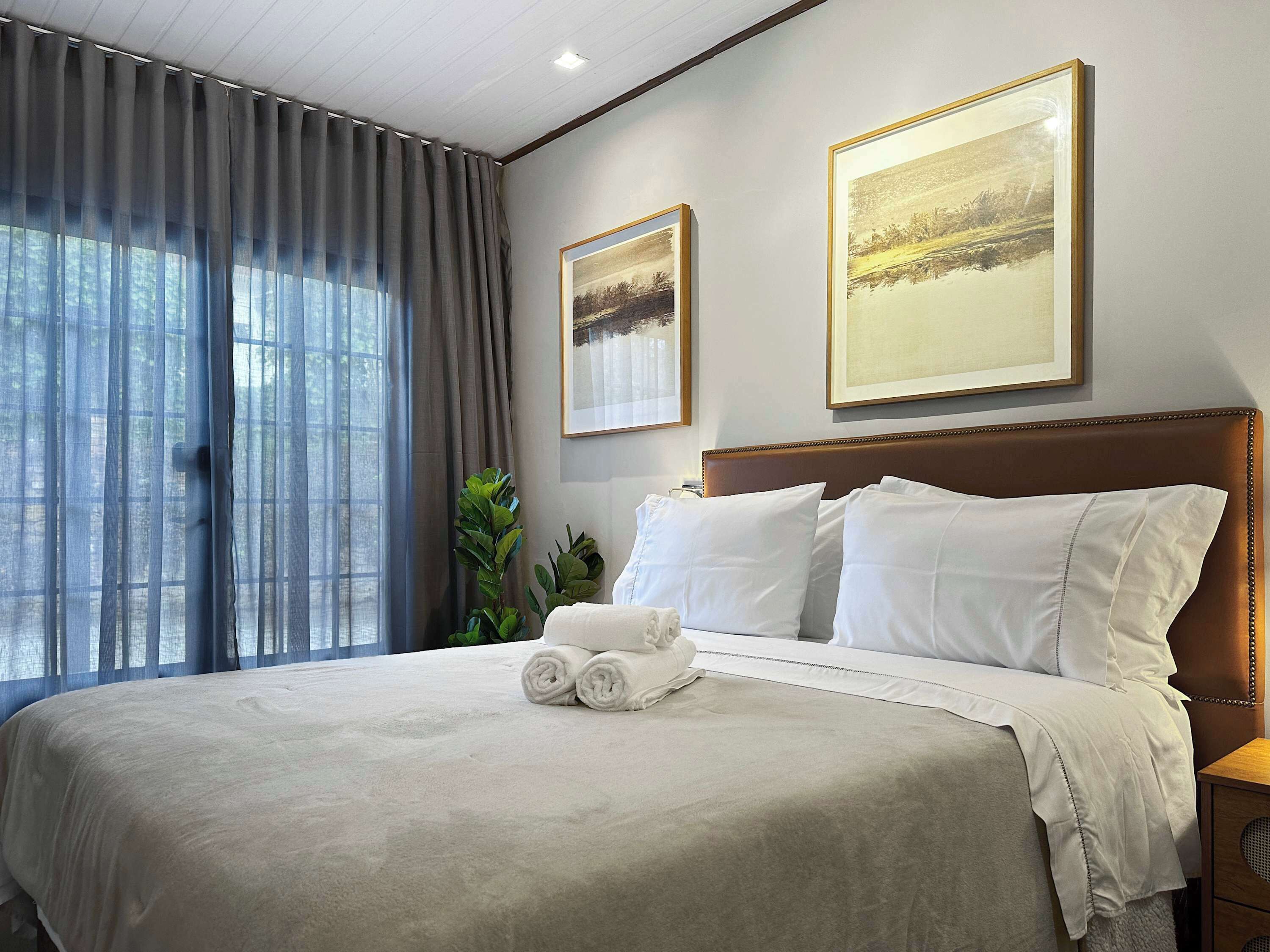 A hotel room with a large bed, two framed pictures on the wall, a window with curtains, and a bedside table.