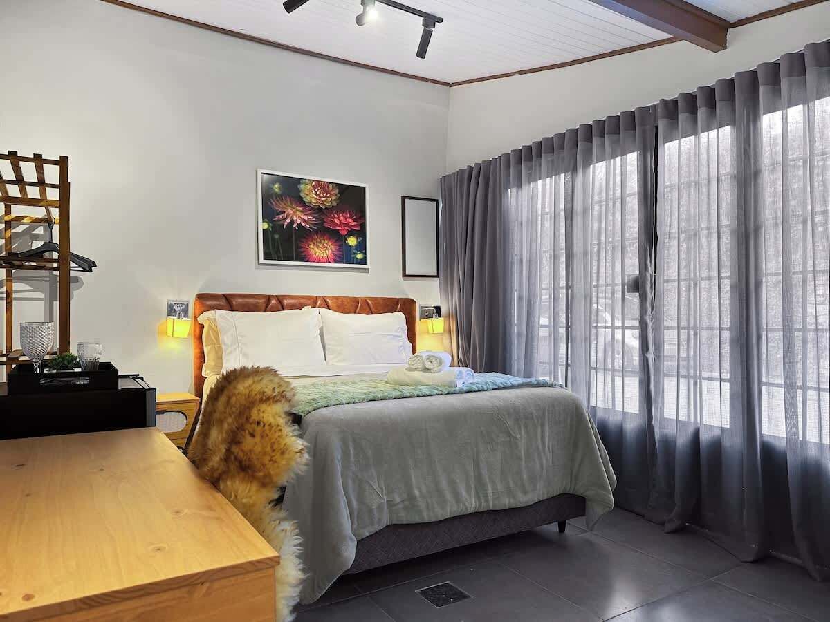 A hotel room with a bed, a wooden desk, a chair, a painting on the wall, and a window with curtains.