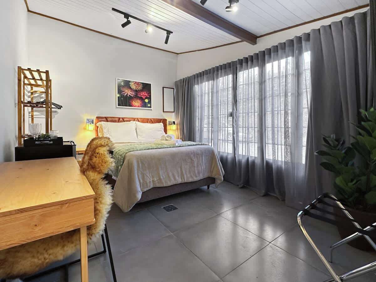 Un dormitorio con cama, escritorio, silla, planta y ventana con cortinas.