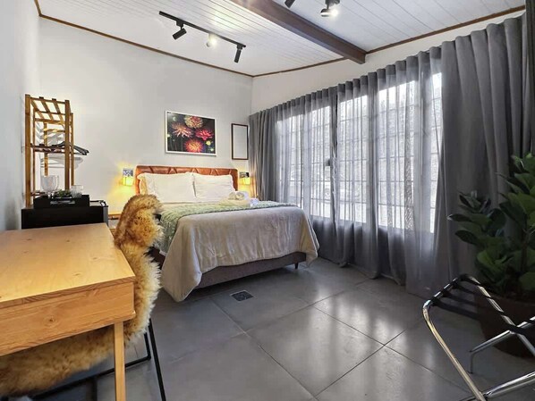 A bedroom with a bed, a desk, a chair, a plant, and a window with curtains.