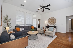 A spacious living room with a grey sofa, a round coffee table, and a ceiling fan. - Steps from Main street, Updated Family-Friendly Home with Fireplace and Garage! (Frisco)