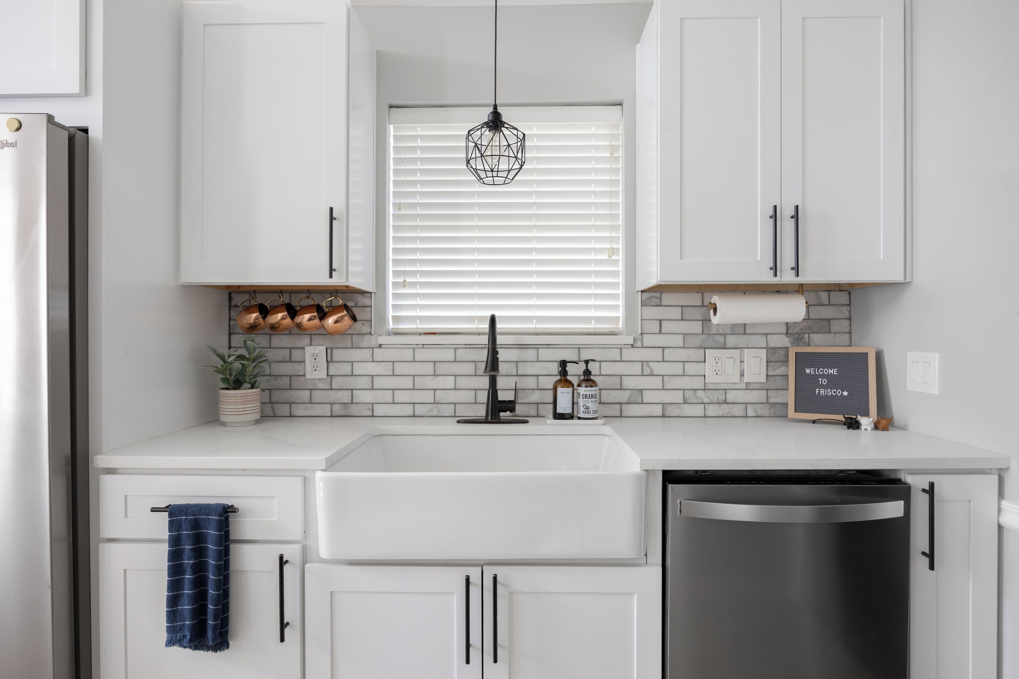 A modern kitchen with white cabinets, a stainless steel dishwasher, and a window with blinds.